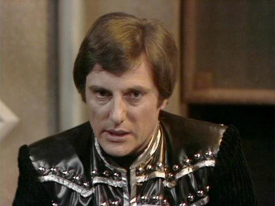 Claude 3.7 describes the image as: "The image shows a person in what appears to be a costume from a science fiction television series. They're wearing a distinctive black outfit with metallic or reflective shoulder elements that have studs or decorative elements. The costume has an elaborate design typical of 1970s-80s British sci-fi productions.

The setting appears to be an indoor studio set with muted colors in the background. The theatrical costume with its dark color scheme and metallic ac…