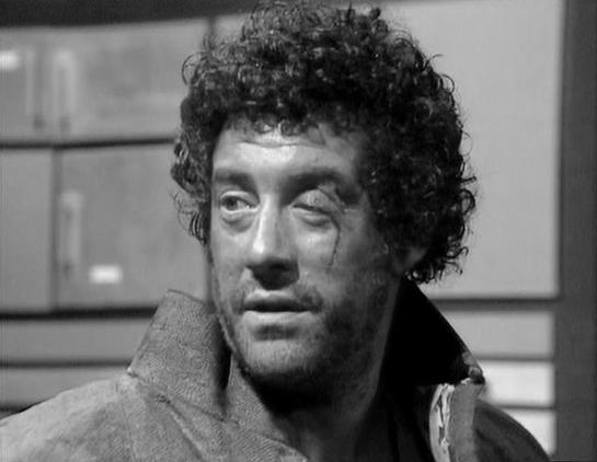 Claude 3.7 describes the image as: "This is a black and white image from a classic British science fiction television series from the late 1970s/early 1980s. The image shows a person with curly, dark hair and a somewhat disheveled appearance, wearing what appears to be a rough-textured coat or jacket with an upturned collar. The expression on their face appears somewhat intense or thoughtful.

The setting seems to be some kind of industrial or spacecraft interior, with what looks like storage c…