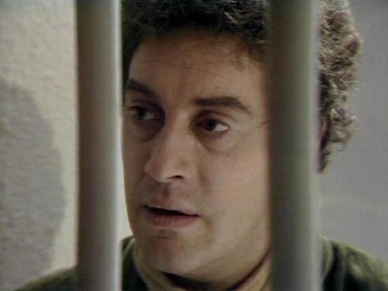Claude 3.7 describes the image as: "The image shows a person behind what appears to be prison bars or some type of confinement. They have curly dark hair and are wearing what looks like an olive or greenish shirt. The scene has a dramatic quality with the bars creating a stark visual separation between the subject and the viewer. The lighting creates shadows that emphasize the confined setting, giving the image a tense, dramatic atmosphere typical of classic science fiction television productio…