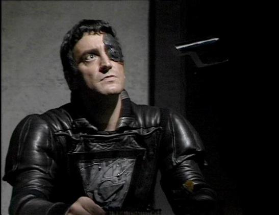 Claude Sonnet 4.0 describes the image as: "I can see this appears to be from a science fiction television production, showing someone in dark, futuristic military-style armor or uniform. The costume design has a distinctive sci-fi aesthetic with metallic elements and structured plating typical of space opera productions from that era. The lighting and set design create a dramatic, somewhat ominous atmosphere with dark tones and institutional-looking backgrounds. The overall production values an…