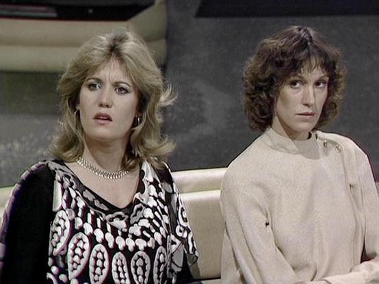 Claude 3.7 describes the image as: "The image shows two people seated together in what appears to be a sci-fi television series set from the late 1970s or early 1980s. The person on the left has blonde layered hair and is wearing a patterned black and white top with a pearl necklace. The person on the right has curly brown hair and is wearing a light-colored top with some subtle detailing at the shoulder.

The styling, lighting, and overall aesthetic are characteristic of British television pro…