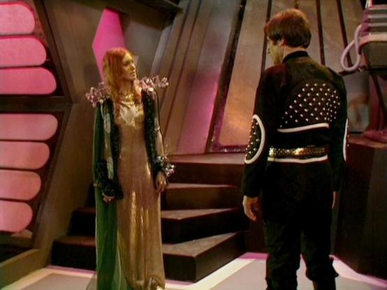 Claude 3.7 describes the image as: "This image appears to be from a science fiction television series, featuring a scene set inside what looks like a futuristic spacecraft or space station. The interior has distinctive angular architecture with pink/reddish lighting elements and metallic walls.

Two figures are shown in conversation. On the left is a person wearing an elaborate costume consisting of a golden/beige dress with a green robe or cloak. On the right is a person dressed in black attir…