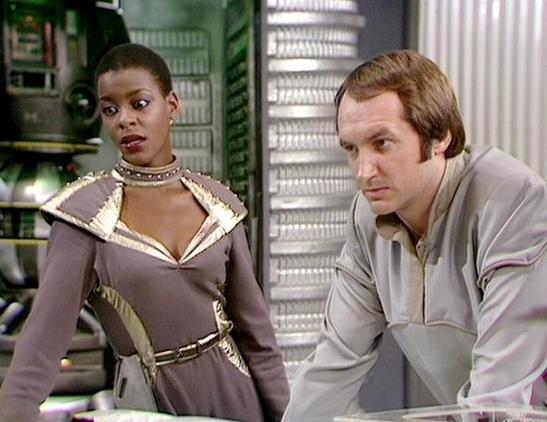 Claude Sonnet 4.0 describes the image as: "This appears to be a scene from a science fiction television series, showing two characters in what looks like a spacecraft or futuristic facility interior. The setting features typical sci-fi production design with metallic walls and technical equipment visible in the background. One character is wearing an elaborate costume with metallic trim and ornate detailing around the collar and belt, while the other is dressed in more utilitarian clothing. The…