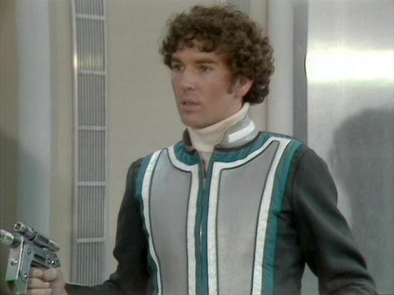 Claude 3.7 describes the image as: "This image shows a scene from what appears to be a science fiction television production from the late 1970s or early 1980s, based on the visual style and production quality. 

The person in the frame is wearing a distinctive futuristic costume with a gray and teal vest-like garment over a dark jacket, with a white turtleneck underneath. They have curly dark hair and are standing in what looks like a spacecraft or space station interior with metallic walls an…