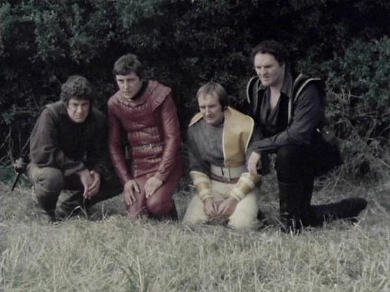 Claude 3.7 describes the image as: "This image appears to be from a vintage science fiction television series from the late 1970s or early 1980s. The scene shows four people in distinctive costume attire kneeling or sitting in a grassy outdoor setting with vegetation in the background.

The costumes are quite notable - one person wears a burgundy/red padded outfit, another is in a beige/yellow costume, while the others are dressed in darker clothing. The styling and production aesthetic is char…