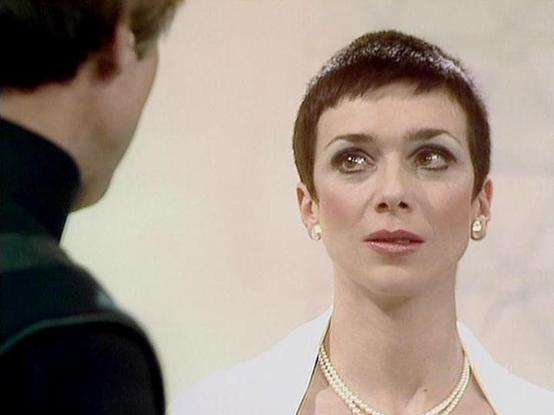Claude Sonnet 4.0 describes the image as: "I can see this appears to be from a science fiction television series, showing what looks like a dramatic scene between two characters. The image shows a person with very short dark hair wearing what appears to be a white garment and pearl necklace, engaged in conversation with someone whose back is to the camera. The setting appears to be indoors with neutral-colored walls. The lighting and film quality suggest this is from a television production, li…