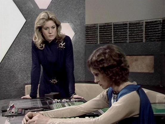 Claude Sonnet 4.0 describes the image as: "This scene takes place in what appears to be the computer room or control center of the Liberator spacecraft. The setting features the show's characteristic futuristic design with geometric wall panels and control surfaces. One woman stands wearing a dark blue outfit with decorative elements, positioned behind another woman who is seated at a control console. The seated figure wears a cream-colored top with blue trim and has shorter, curled hair. The s…