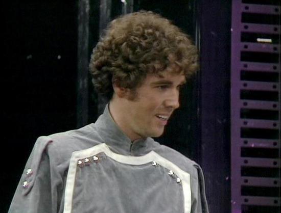 Claude Sonnet 4.0 describes the image as: "I can see this is a scene featuring a character in a distinctive futuristic gray and white uniform with metallic studs or buttons along the chest. The costume has a military or space-faring aesthetic typical of science fiction television from this era. The setting appears to be aboard a spacecraft or space station, with dark metallic walls and what looks like technical equipment or control panels visible in the background. The lighting creates a dramat…