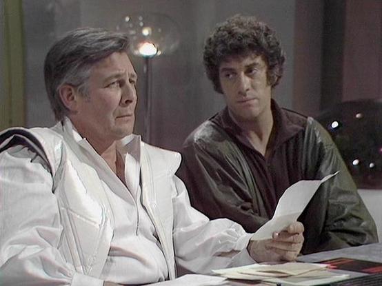 Claude 3.7 describes the image as: "This image appears to be from a classic British science fiction television series from the late 1970s/early 1980s. The scene shows two men in what looks like a futuristic or institutional setting. On the left is an older man wearing a white uniform or outfit, possibly representing authority or medical profession, holding what appears to be a document or paper. To his right is a younger man with curly dark hair wearing a dark shirt or jacket, who seems to be l…