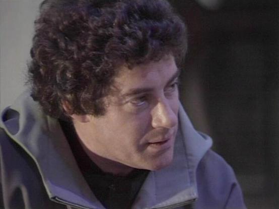 Claude Sonnet 4.0 describes the image as: "I can see this is a scene featuring a man with dark curly hair wearing what appears to be a light-colored, futuristic-style jacket or uniform. The lighting and composition suggest this is from a science fiction television production, likely from the late 1970s or early 1980s based on the visual style and production values. The setting appears to be indoors, possibly on a spacecraft or futuristic facility set. The character appears to be in conversation…