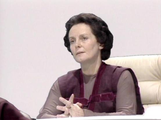Claude 3.7 describes the image as: "The image shows a person in what appears to be a formal setting, possibly from a science fiction television production from the late 1970s or early 1980s based on the visual quality and styling. 

The individual is wearing a distinctive burgundy vest or sleeveless garment over a lighter colored top, and is seated in what looks like an official capacity, possibly at some kind of control panel or council table. They appear to be in a white or light-colored chai…