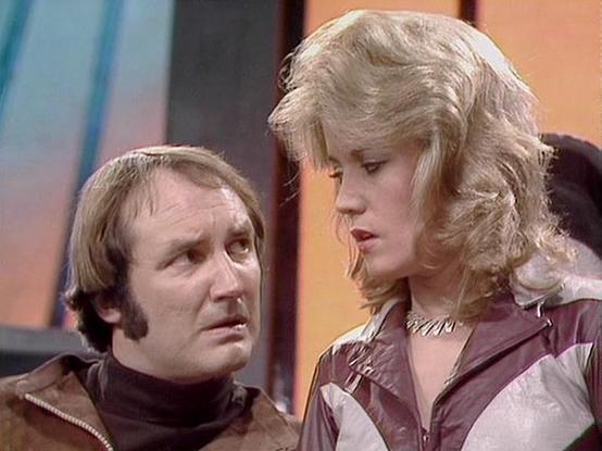 Claude Sonnet 4.0 describes the image as: "I can see two people in what appears to be a science fiction television setting, likely aboard a spacecraft based on the futuristic interior visible in the background. One person has shoulder-length blonde hair and is wearing what looks like a burgundy or maroon colored outfit with metallic detailing. The other person has darker hair and is wearing what appears to be a brown leather or similar material jacket. They seem to be engaged in conversation, w…