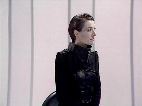 Claude 3.7 describes the image as: "This image appears to be from a science fiction television production, featuring a person in a sleek black uniform with a high collar and military-style design. The setting has a minimalist, futuristic aesthetic with vertical white panels forming the background, giving it a sterile, possibly spaceship or high-tech facility appearance. The subject is shown in profile, wearing their dark hair slicked back, which complements the austere, utilitarian style of the…