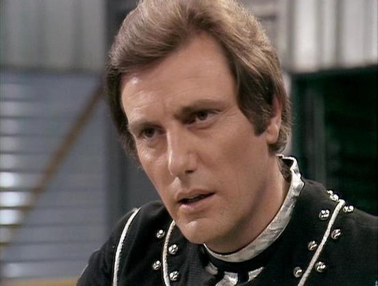 Claude 3.7 describes the image as: "The image shows a person in what appears to be a science fiction setting. They are wearing a distinctive black costume with metallic embellishments or studs along the collar area, and a light-colored undershirt or collar visible beneath. The styling has a classic vintage sci-fi aesthetic typical of British television productions from the late 1970s or early 1980s.

The scene appears to be set in some kind of spacecraft or futuristic facility, with stairs visi…