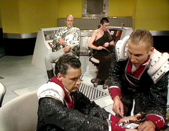Claude Sonnet 4.0 describes the image as: "This scene appears to be set on a futuristic spacecraft bridge or control room, characterized by the sleek white and metallic control panels and the distinctive yellow and black striped walls typical of the series' production design. The setting has that clinical, high-tech aesthetic common to science fiction television of the era.

The scene shows four characters in elaborate costumes. Two figures in the foreground are wearing ornate black and white o…