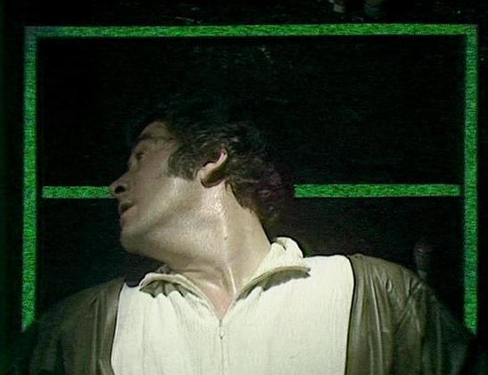 Claude Sonnet 4.0 describes the image as: "I can see this appears to be from a science fiction television production, showing a person in what looks like a white or light-colored shirt or jacket against a dark background with green geometric framing elements. The lighting and visual style is characteristic of 1970s or early 1980s British television production. The setting appears to be some kind of interior space, possibly a spacecraft or futuristic facility, with the distinctive green-tinted g…