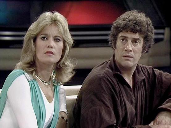 Claude 3.7 describes the image as: "This image appears to be from a science fiction television series from the late 1970s or early 1980s. It shows two people in what looks like a spacecraft or futuristic control room setting. On the left is a person with blonde feathered hair wearing a white outfit with a teal/mint green accent and a gold chain necklace. On the right is a person with dark curly hair wearing a brown shirt. Both have serious or concerned expressions as they appear to be reacting …