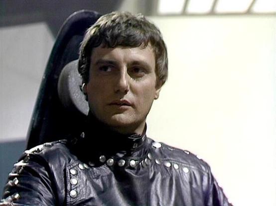 Claude 3.7 describes the image as: "The image shows a person in what appears to be a science fiction setting, wearing a distinctive dark leather or vinyl costume with metallic studs or decorative elements. They are seated in what looks like a high-backed chair or control station with light-colored padding visible behind them. The costume has a futuristic, somewhat militaristic style typical of classic science fiction television productions from the late 1970s or early 1980s. The lighting and fi…