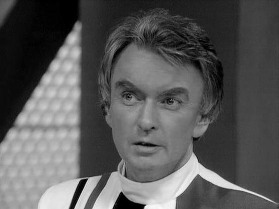 Claude 3.7 describes the image as: "This is a black and white image from a science fiction television production. The person in the image is wearing what appears to be a distinctive uniform or costume with a white collar and dark suspender-like elements. The setting seems to be a futuristic or spacecraft interior, with some geometric patterns visible in the background.

The image has the characteristic aesthetic of British science fiction television from the late 1970s or early 1980s, with mini…