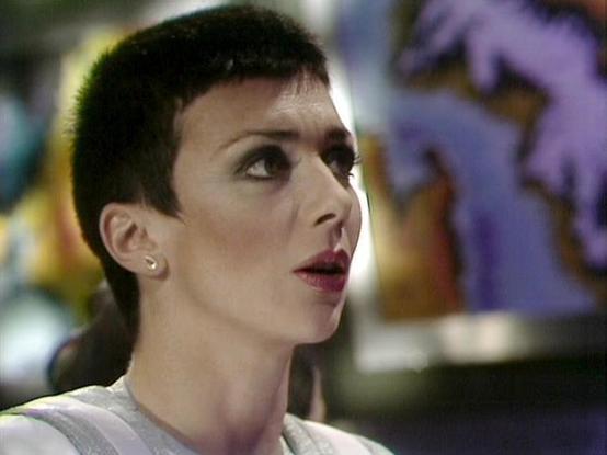 Claude Sonnet 4.0 describes the image as: "I can see this appears to be from a science fiction television production, likely from the late 1970s or early 1980s based on the visual style and production quality. The image shows a person with very short, dark hair and distinctive makeup, wearing what appears to be a light-colored garment. The setting appears to be some kind of futuristic or sci-fi environment with blurred lighting effects in the background. The cinematography and costume design ar…