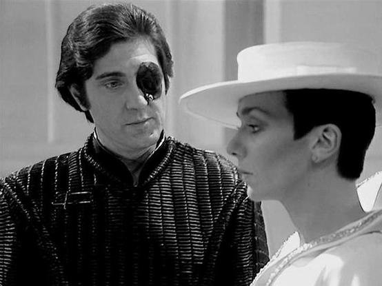 Claude Sonnet 4.0 describes the image as: "I can see this is a black and white scene showing two characters in what appears to be a dramatic confrontation. One figure wears an ornate, metallic costume with distinctive armor-like detailing, while the other is dressed in white with a wide-brimmed hat. The setting appears to be an interior space with clean, futuristic architecture typical of science fiction productions. The composition and lighting suggest this is a pivotal dramatic moment between…