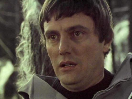 Claude 3.7 describes the image as: "The image shows a close-up of a person with dark, short hair wearing what appears to be a gray or silver jacket with a high collar. They have an intense or concerned expression on their face. The lighting gives the scene a somewhat dark, muted tone with greenish hues in the background.

This appears to be from a science fiction television production from the late 1970s or early 1980s, based on the visual style and filming quality. The setting seems to be some…