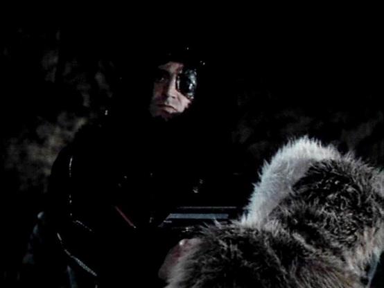 Claude 3.7 describes the image as: "The image shows a dark, atmospheric scene featuring a figure in what appears to be a black leather outfit with some kind of eye patch or facial covering. They are partially illuminated against a very dark background, creating a dramatic, shadowy effect. The costume includes what looks like fur trim visible in the lower right portion of the frame.

This appears to be from a science fiction production, with the styling and costume design suggesting a dystopian …