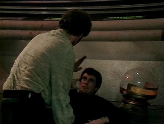 Claude 3.7 describes the image as: "The image shows a scene from what appears to be a science fiction television production from the late 1970s or early 1980s, based on the visual style and production quality. 

The setting looks like the interior of a spacecraft or space station with curved walls and minimalist design elements typical of sci-fi of that era. Two people are visible in the frame - one person in a light-colored shirt standing and gesturing to another person wearing dark clothing w…