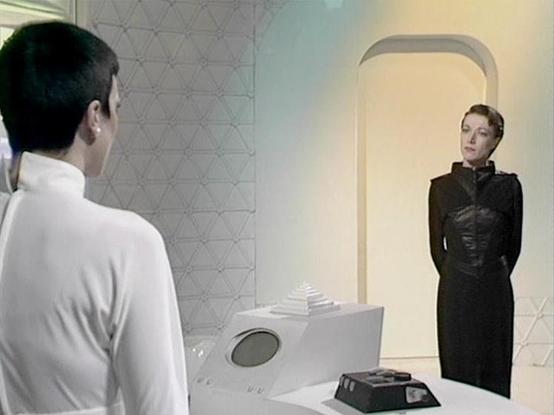 Claude 3.7 describes the image as: "This image appears to be from a science fiction television series, showing a scene in a minimalist, futuristic setting with white walls featuring a geometric pattern. The scene depicts two people in a formal conversation across what appears to be a control console or technical device with buttons and a circular display.

On the left is a person with short dark hair wearing a white outfit, shown from behind. On the right stands someone in a fitted black leathe…