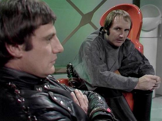 Claude Sonnet 4.0 describes the image as: "This appears to be a scene from a science fiction television series, showing two characters in what looks like a spacecraft or futuristic setting. One character is wearing a distinctive black leather outfit with metallic studs or decorative elements, while the other is dressed in more casual clothing and appears to be wearing headphones or some kind of communication device. The setting has a retro-futuristic aesthetic typical of British sci-fi producti…