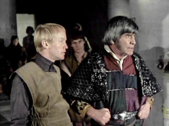 Claude Sonnet 4.0 describes the image as: "I can see this appears to be from a science fiction television production, showing two men in what looks like a futuristic or medieval-style setting. One man has blonde hair and is wearing what appears to be a light-colored vest or tunic, while the other has darker hair and is dressed in more elaborate clothing with metallic or chainmail-like material. The setting appears to be indoors with other figures visible in the background. The lighting and prod…