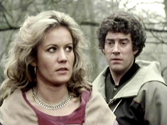 Claude Sonnet 4.0 describes the image as: "I can see this appears to be from a science fiction television series, showing two people in what looks like an outdoor setting with trees in the background. The image has the characteristic film quality and styling typical of British television from the late 1970s or early 1980s. One person is wearing what appears to be a burgundy or wine-colored top with a metallic chain necklace, while the other is dressed in what looks like a light-colored jacket o…