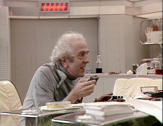 Claude Sonnet 4.0 describes the image as: "I can see this is a scene set in what appears to be a futuristic medical facility or laboratory, with clean white walls and advanced-looking equipment visible in the background. The setting has the distinctive sterile, high-tech aesthetic typical of science fiction productions from this era. An elderly man with grey curly hair is seated at what looks like a examination or work table, wearing casual clothing. The scene appears to be from a dramatic mome…