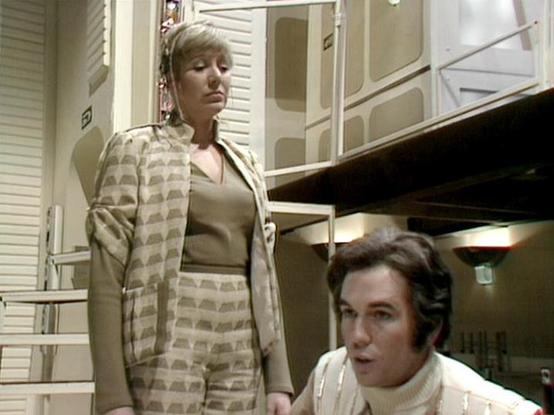 Claude 3.7 describes the image as: "The image shows a scene from what appears to be a retro sci-fi production, featuring two people in a futuristic interior setting. One person is standing wearing a distinctive patterned outfit in neutral tones - a matching jacket and pants with a geometric pattern, paired with a light-colored top. The other person is seated below, wearing what looks like a cream-colored turtleneck sweater.

The setting has a very distinctive 1970s-era science fiction aesthetic…