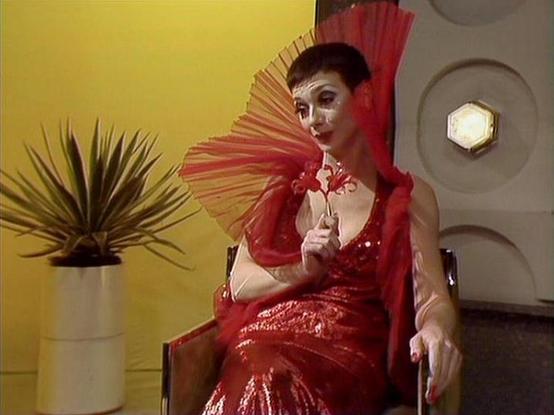 Claude 3.7 describes the image as: "The image shows a performer in a striking red sequined dress with a dramatic fan-like collar or accessory in matching red. Their theatrical makeup includes distinctive facial cosmetics that create a bold, stylized appearance. They're seated in what appears to be a retro-futuristic setting with a yellow wall, a potted plant, and geometric wall features including a hexagonal light fixture.

The scene has a theatrical, dramatic quality with the vibrant red costu…