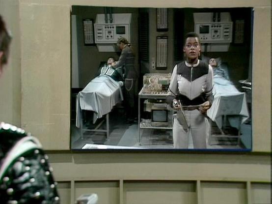 Claude 3.7 describes the image as: "This image appears to be from a science fiction television series, showing what looks like a medical or technical facility. The scene features several people in a clinical setting with control panels and equipment visible in the background. 

In the foreground, a person wearing a distinctive black and white uniform stands holding what appears to be a device or tool. Behind them are beds or examination tables with people lying on them covered by white sheets. …