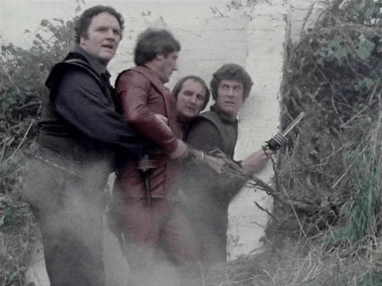 Claude Sonnet 4.0 describes the image as: "I can see four men in what appears to be an outdoor, rocky or cave-like setting with vegetation around them. They're wearing what looks like futuristic or science fiction-style clothing in muted colors - dark and earth tones. The scene has a gritty, dramatic quality typical of 1970s science fiction television production. The lighting and atmosphere suggest this might be during an action sequence or tense moment in the story. The setting appears rough a…