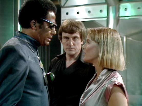 Claude 3.7 describes the image as: "This image appears to be from a science fiction television series from the late 1970s or early 1980s. The scene shows three people in what looks like a futuristic spacecraft or space station interior with metallic walls and distinctive lighting.

The image captures a tense conversation between three characters. On the left is a person wearing glasses and a uniform with what appears to be a badge or emblem. In the center is someone dressed in black attire, loo…
