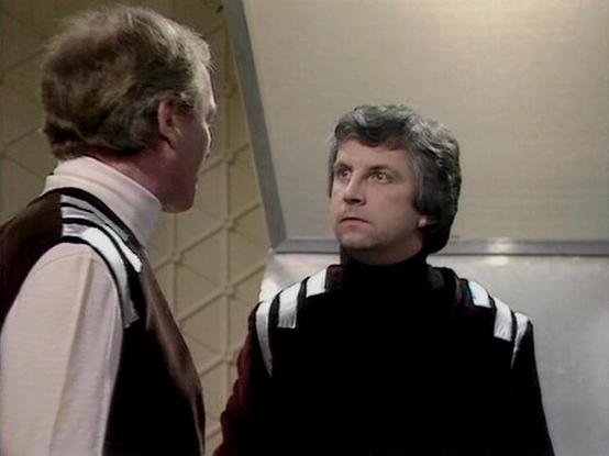 Claude 3.7 describes the image as: "This image appears to be from a classic science fiction television series from the late 1970s/early 1980s. The scene shows two people in a conversation, set in what looks like a futuristic spacecraft or space station interior with geometric wall patterns typical of sci-fi productions from that era.

The two individuals are wearing distinctive costumes with unusual collar designs - one in a dark outfit with striped shoulder details, and the other in lighter at…
