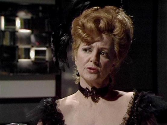 Claude Sonnet 4.0 describes the image as: "I can see this is a scene from a science fiction television series featuring an elegantly dressed woman in what appears to be a Victorian or period-inspired costume. The character is wearing an ornate black dress with decorative details and a matching choker necklace, with her blonde hair styled in an elaborate updo typical of 19th century fashion. The setting appears to be aboard a spaceship or futuristic facility, with modern lighting and metallic su…