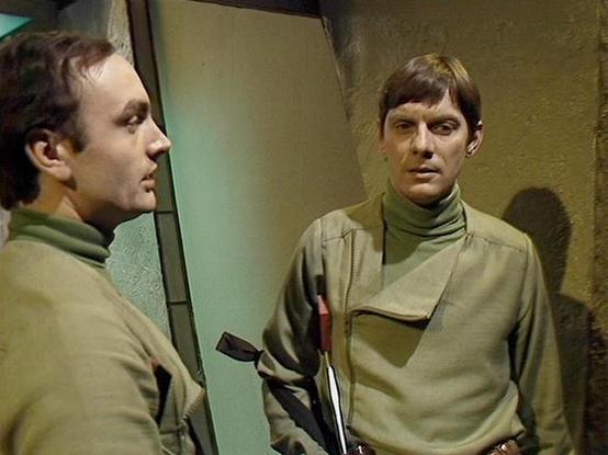 Claude 3.7 describes the image as: "This image appears to be from a vintage science fiction television production, likely from the late 1970s or early 1980s based on the visual style and filming quality. 

The scene shows two individuals in a sparse, utilitarian setting with plain walls. Both are wearing similar olive-green high-necked uniforms or jumpsuits that have a military or institutional appearance. One person is visible in profile on the left, while another is facing forward on the righ…