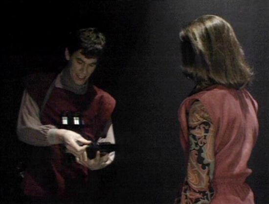 Claude 3.7 describes the image as: "The image shows two people in what appears to be a dark setting. On the left is a person wearing a burgundy vest over a light-colored shirt, who appears to be holding or examining some kind of device or object. On the right is someone with shoulder-length hair wearing a pink or salmon-colored outfit with decorative patterned details.

The scene has a vintage science fiction aesthetic with the dark background creating a dramatic atmosphere. The lighting is low…