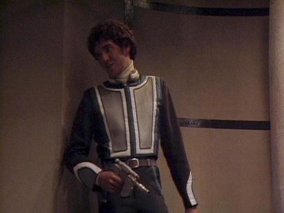 Claude 3.7 describes the image as: "This image shows a person in what appears to be a science fiction costume or uniform. They're wearing a distinctive outfit with a gray/black base and cream-colored panels on the front, with contrasting trim and a belt. The person is holding what looks like a prop weapon or futuristic gun. 

The setting appears to be a minimalist interior with plain walls, typical of science fiction television productions from the late 1970s or early 1980s based on the visual …