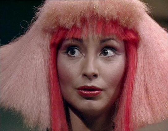 Claude Sonnet 4.0 describes the image as: "I can see this is a science fiction television scene featuring a character with distinctive styling - voluminous blonde and pink/red hair, and dramatic makeup including red lipstick. The character appears to be in some kind of futuristic or otherworldly setting, which is typical of the sci-fi genre. The styling and production values suggest this is from a British science fiction series from the late 1970s or early 1980s era. However, I cannot identify …
