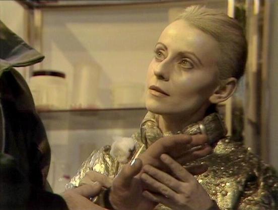 Claude Sonnet 4.0 describes the image as: "I can see this appears to be from a science fiction television production, showing what looks like a dramatic scene in a futuristic or clinical setting. The image shows a person with pale, almost ethereal makeup and styling, wearing what appears to be a metallic or reflective costume. The lighting and set design suggest this is from a studio production, with the characteristic look of British science fiction television from that era. The scene appears …
