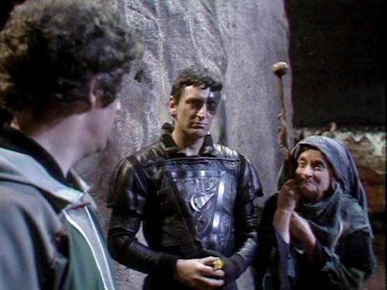 Claude Sonnet 4.0 describes the image as: "I can see this appears to be from a science fiction television production, showing what looks like a dramatic scene in what appears to be a cave or rocky underground setting. There are several people in the scene - one person wearing what appears to be futuristic armor or protective gear with a metallic chest piece, and others in darker clothing. The lighting and set design has that distinctive look of British science fiction television from the late 1…