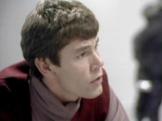 Claude 3.7 describes the image as: "The image shows a person in a side profile view with short dark hair. They appear to be wearing a burgundy or maroon colored outfit with what looks like a lighter colored collar or scarf. The setting appears to be from a television production, with a somewhat muted color palette typical of productions from the late 1970s or early 1980s.

The composition suggests this is from a dramatic scene, with the subject looking intently at something off-camera. The ligh…