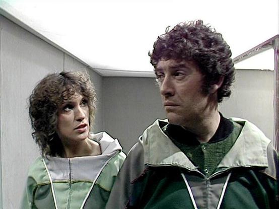 Claude Sonnet 4.0 describes the image as: "I can see two people in what appears to be a futuristic or sci-fi setting, likely aboard a spacecraft or space facility based on the sterile, metallic interior visible in the background. They're wearing matching green and white/silver colored outfits that have a utilitarian, space-age design - possibly uniforms or protective gear. Both individuals have dark, curly hair typical of late 1970s styling. The woman appears concerned or apprehensive, while th…