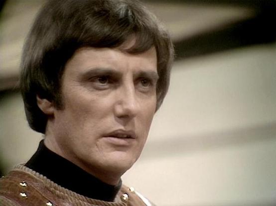 Claude 3.7 describes the image as: "The image shows a person with dark, short hair styled in a typical 1970s-80s fashion. They're wearing what appears to be a high-necked costume with a brown textured material featuring some decorative elements or studs.

The lighting and film quality suggest this is from a British science fiction television production of that era. The costume design has that distinctive retro sci-fi aesthetic common in shows from this period.

The scene appears to be from a st…