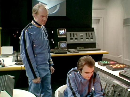 Claude 3.7 describes the image as: "The image shows a scene in what appears to be a control room or spacecraft bridge setting. Two people are wearing similar blue uniforms with distinctive styling that suggests a science fiction production from the late 1970s or early 1980s. 

One person is standing while looking down at the other, who is seated at what looks like a control console with various buttons, lights, and technical equipment. The control room has a futuristic design aesthetic typical …